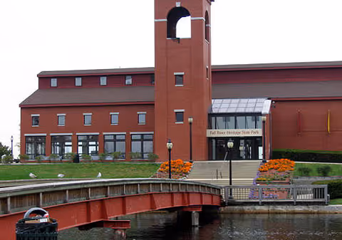Red-brick waterfront building with a central tower, entrance steps, flower beds, and a small red pedestrian bridge over the water in front.