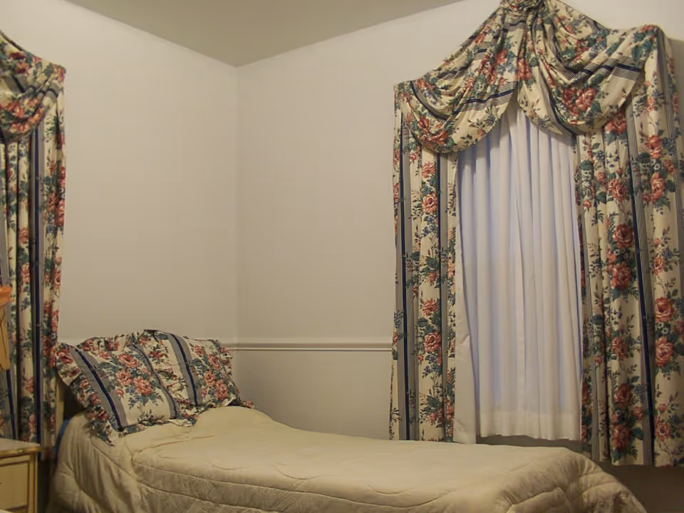A small bedroom with a single bed covered in a beige quilt. The bed has two pillows with floral pillowcases that match the floral curtains on the window beside the bed. The walls are plain and light-colored.