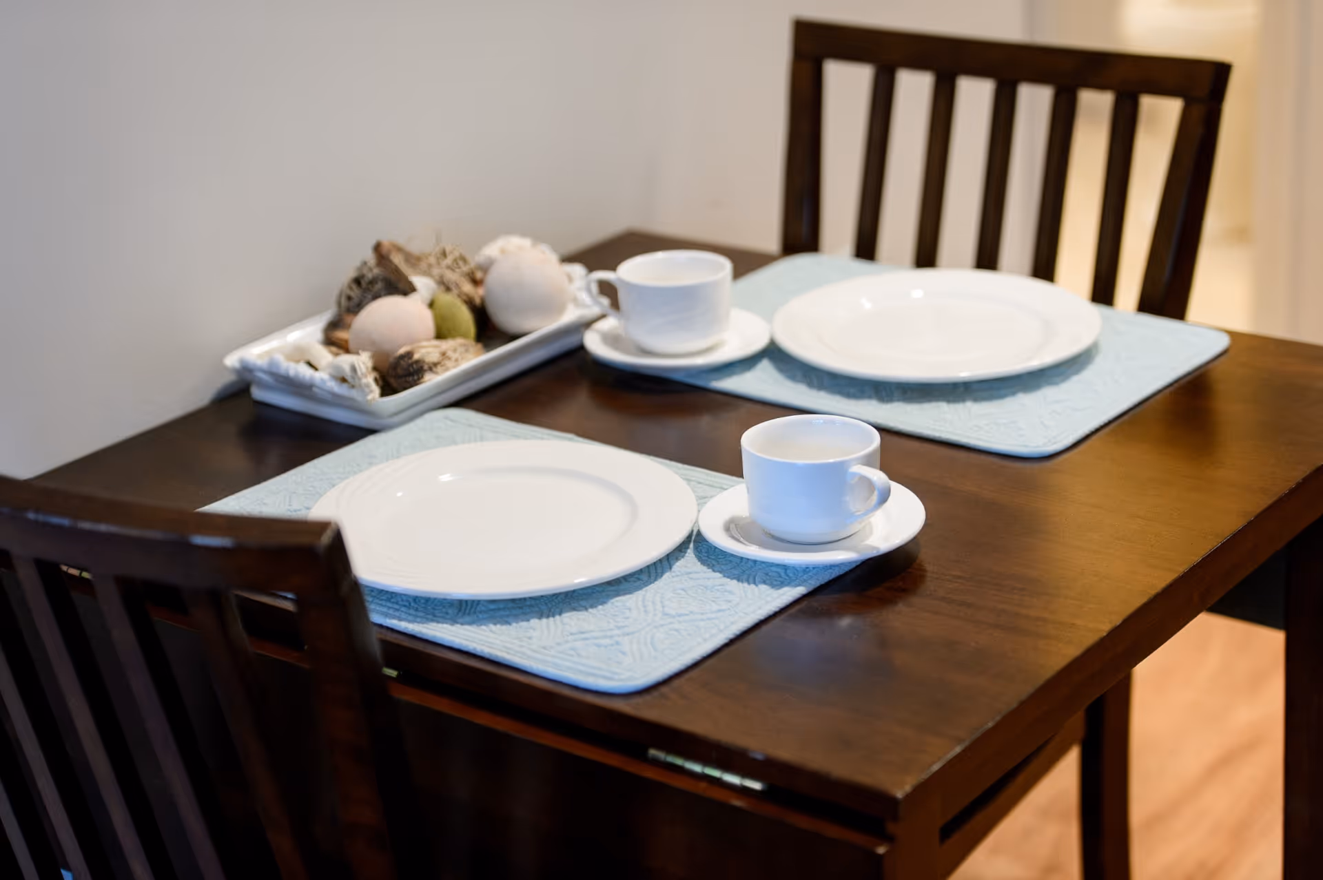 A wooden dining table set for two with light blue placemats, white plates, and white cups with saucers. A rectangular white tray with decorative natural elements is placed on the table. Two wooden chairs with vertical slats are positioned at the table.