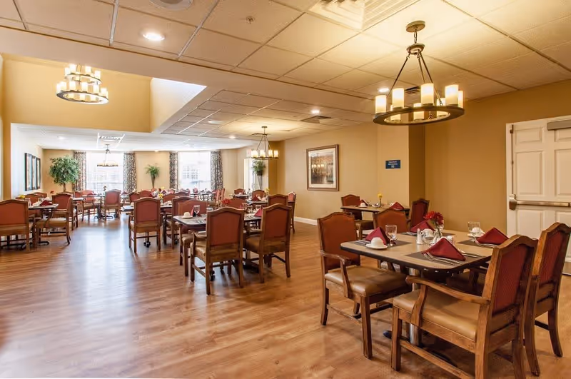 A spacious dining room with multiple wooden tables and chairs arranged neatly. Each table is set with glasses, napkins folded in a triangular shape, and small flower vases. The room has warm lighting from ceiling chandeliers and large windows with curtains allowing natural light to enter. The floor is wooden, and there are framed pictures and plants decorating the walls and corners.