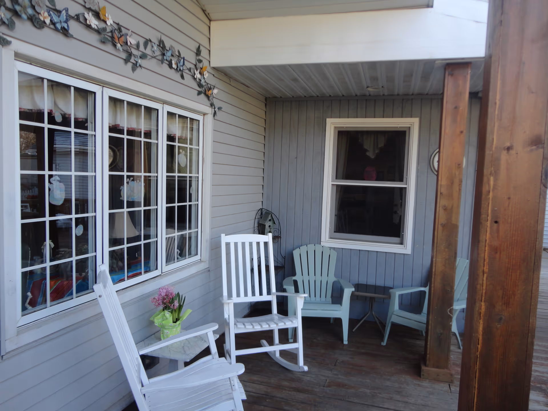 Covered outdoor porch area with wooden floor and ceiling, featuring two white rocking chairs, two light blue plastic chairs, a small table with a potted plant, and decorative butterfly wall art above a window.