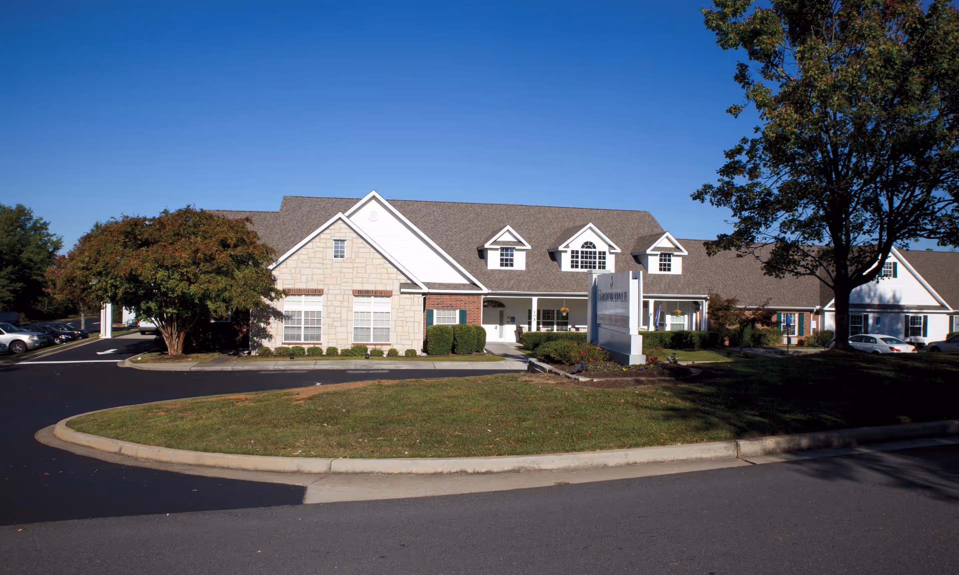 Front exterior of a single-story senior living facility with a lawn, driveway, trees and a sign under a clear blue sky.