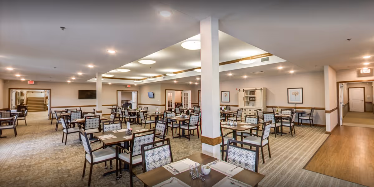 A spacious dining room in a senior living facility with multiple tables and chairs arranged neatly. The room is well-lit with ceiling lights and has neutral-colored walls and carpeted floors. There are framed pictures on the walls and a cabinet with decorative items. Doorways lead to other parts of the facility.