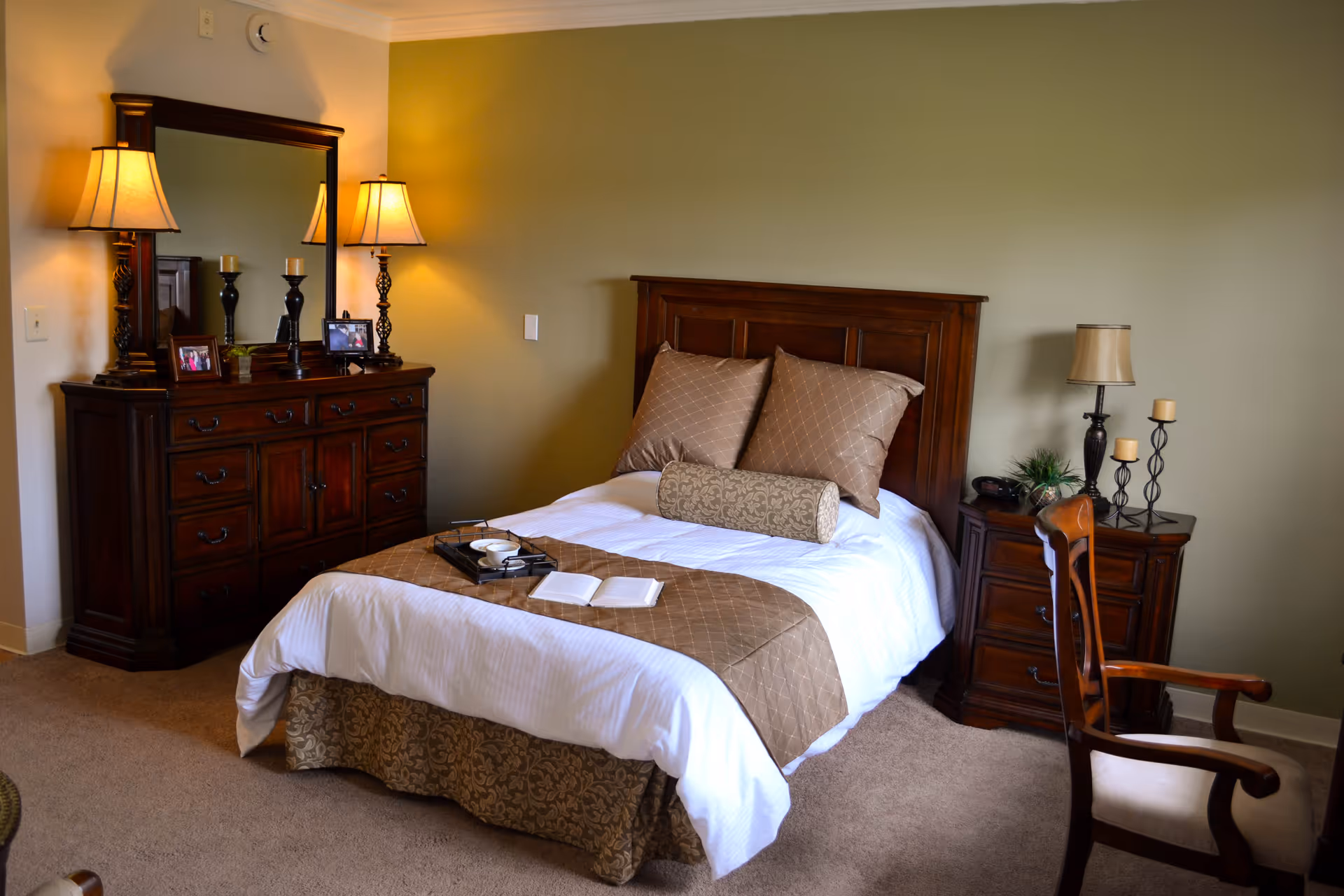 A cozy bedroom with a wooden bed frame, white and brown bedding, two large pillows, and a cylindrical bolster pillow. There is a wooden dresser with a large mirror and two lit table lamps on either side. A wooden nightstand with a lamp, candles, and a small plant is next to the bed. A wooden chair with a cushioned seat is positioned near the nightstand. The walls are painted light green and the floor is carpeted.