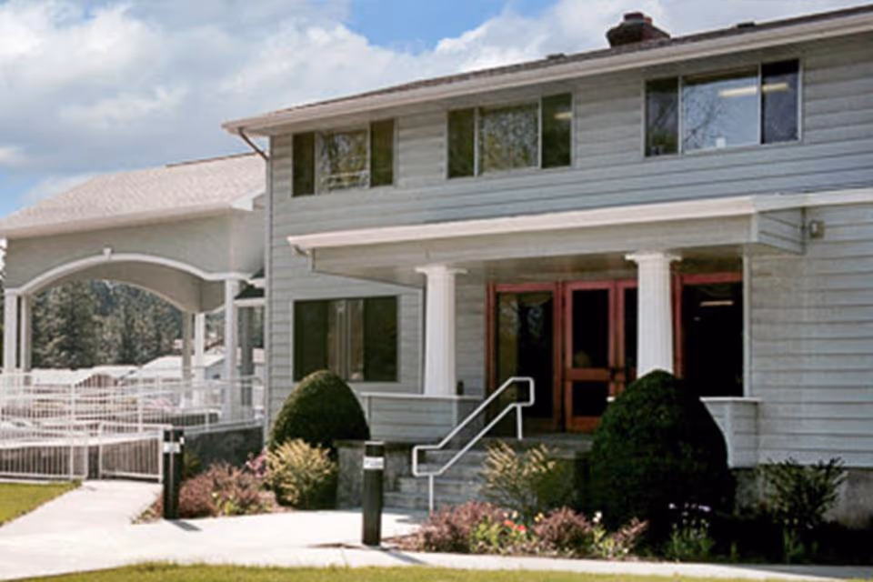 Exterior view of a two-story building with light gray siding and white columns at the entrance. There are bushes and landscaped plants near the entrance steps, and a covered walkway extends from the left side of the building. The sky is partly cloudy.