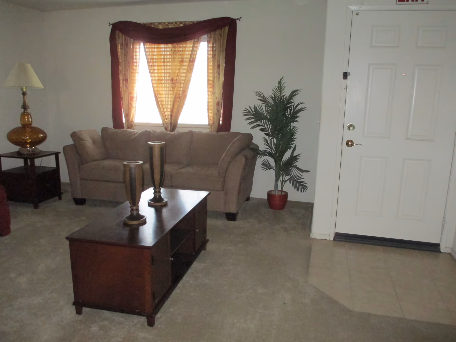 A living room with a beige sofa in front of a window with patterned curtains. There is a wooden coffee table with two decorative vases on it, a side table with a lamp, a potted plant near the white front door, and a carpeted floor transitioning to tiled flooring near the door.