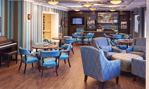 A spacious and well-lit common area with multiple round tables and blue cushioned chairs arranged for dining or socializing. There are also comfortable blue patterned armchairs and a matching ottoman in the foreground. The room features wood flooring, striped wallpaper, and a piano on the left side. In the background, there is a bar area with shelves holding glasses and bottles, and a television mounted on the wall.