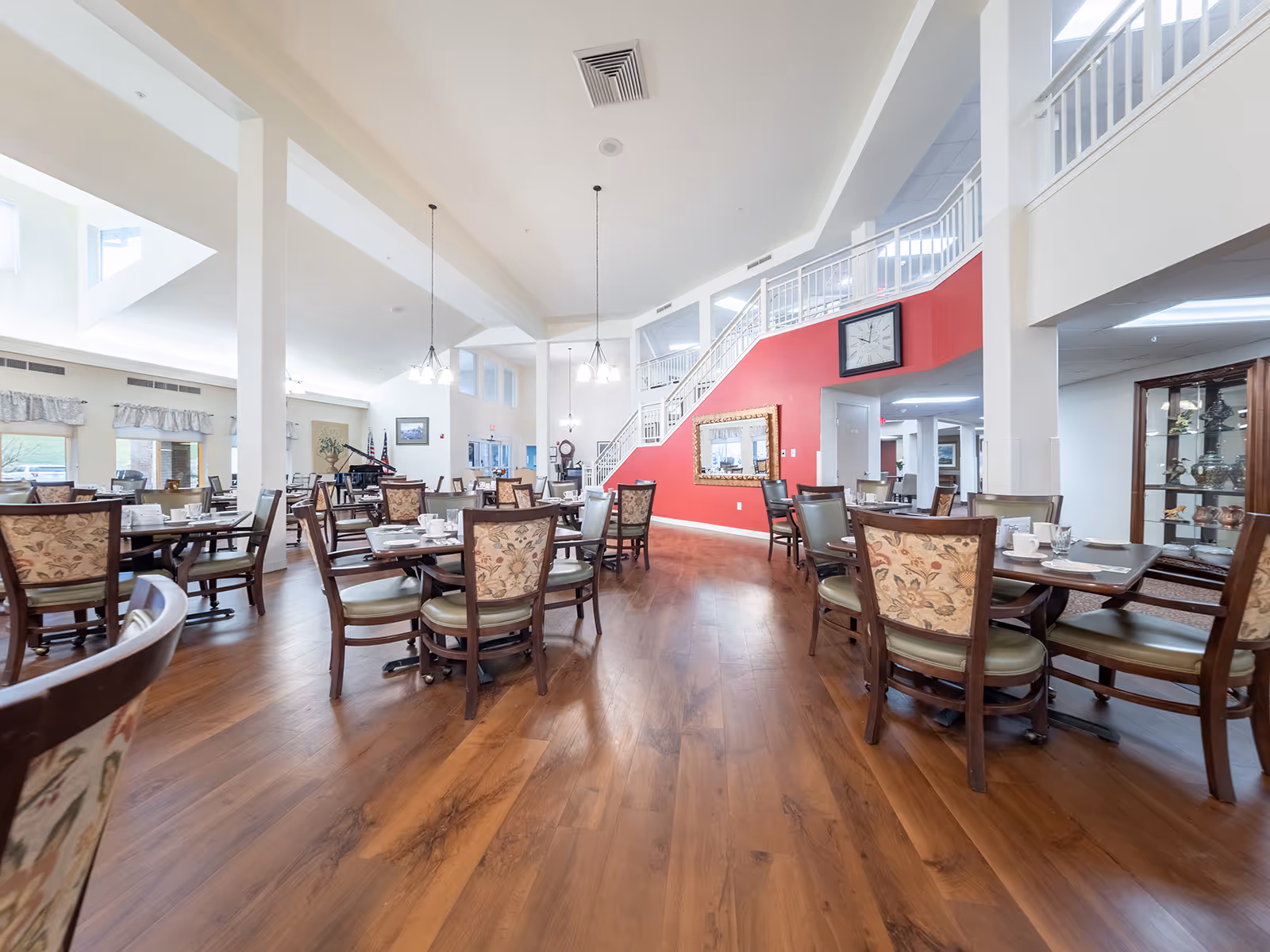 Spacious dining room with wooden floors, multiple set tables and chairs, and a red accent wall with a staircase.