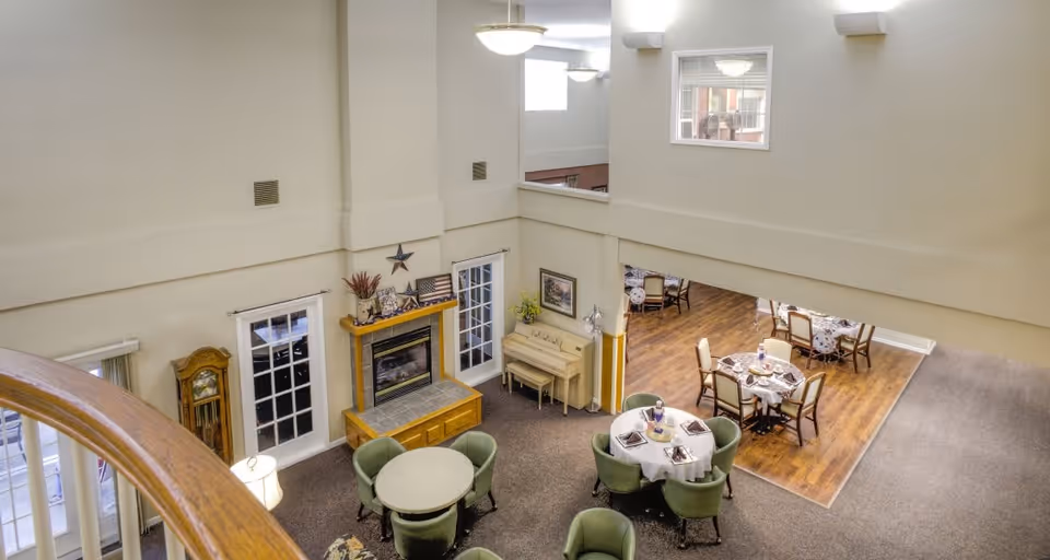 View from an upper level overlooking a common area in a senior living facility. The area features round tables with green chairs, a fireplace with decorative items on the mantel, a piano against the wall, and a dining area with tables and chairs on a wooden floor. The walls are light-colored, and there are windows letting in natural light.