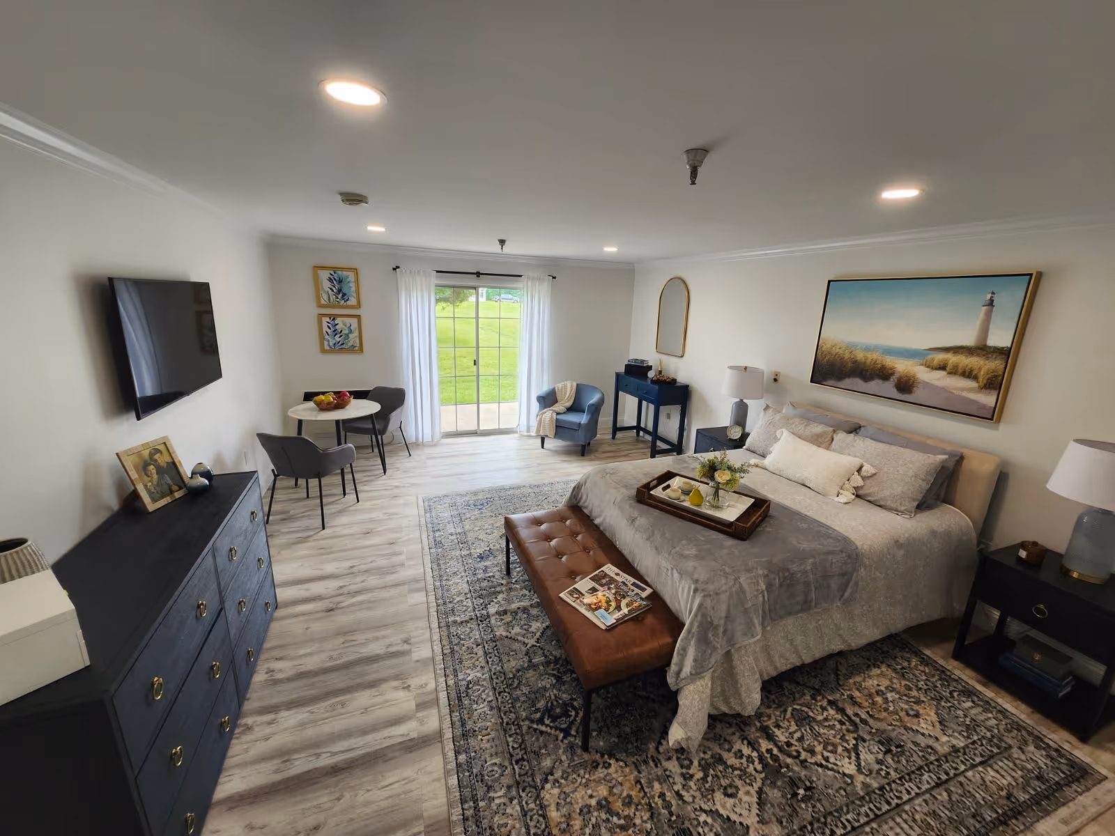 A spacious and well-lit bedroom featuring a large bed with gray bedding and decorative pillows, a brown leather bench at the foot of the bed, a patterned area rug, two nightstands with lamps, a blue dresser with a TV mounted above it, a small round dining table with two chairs, a blue armchair, and a large window with white curtains overlooking a green lawn.