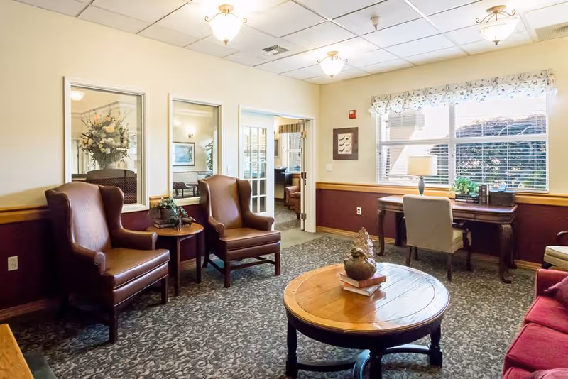 A cozy sitting area in a senior living facility with two brown leather armchairs around a small round wooden table, a round wooden coffee table with a decorative sculpture and books, a desk with a lamp and chair by a window with blinds, and a patterned carpet. The walls are painted cream and maroon with wooden trim, and there are ceiling lights and a doorway leading to another room.