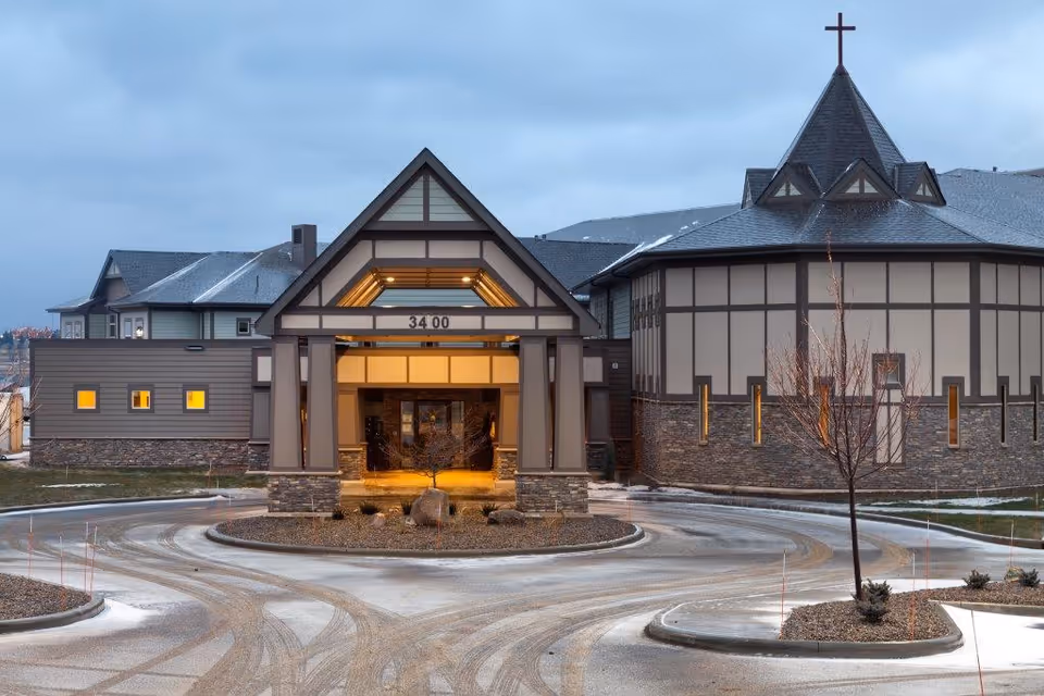 Entrance and circular driveway of a large senior living facility with a peaked portico and a chapel-like wing topped by a cross.