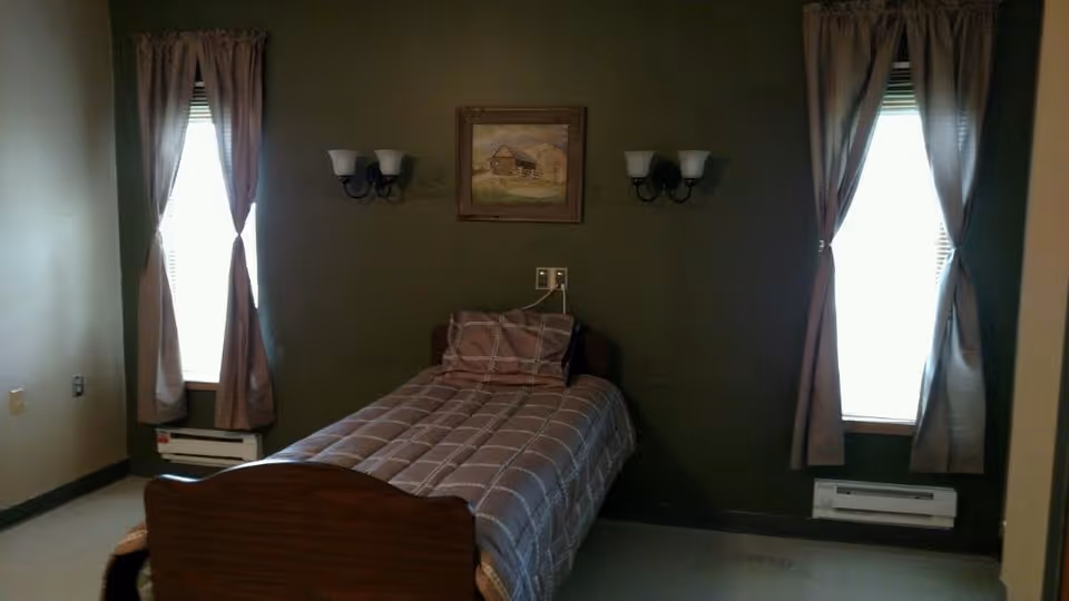 Small bedroom with a single bed against an olive-green wall, two windows with curtains, wall sconces and a framed picture above the bed.