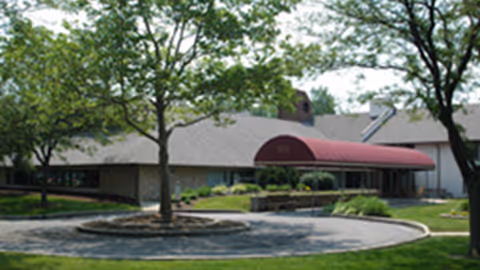 Exterior view of a single-story building with a sloped roof and a red awning over the entrance, surrounded by trees and landscaped greenery.