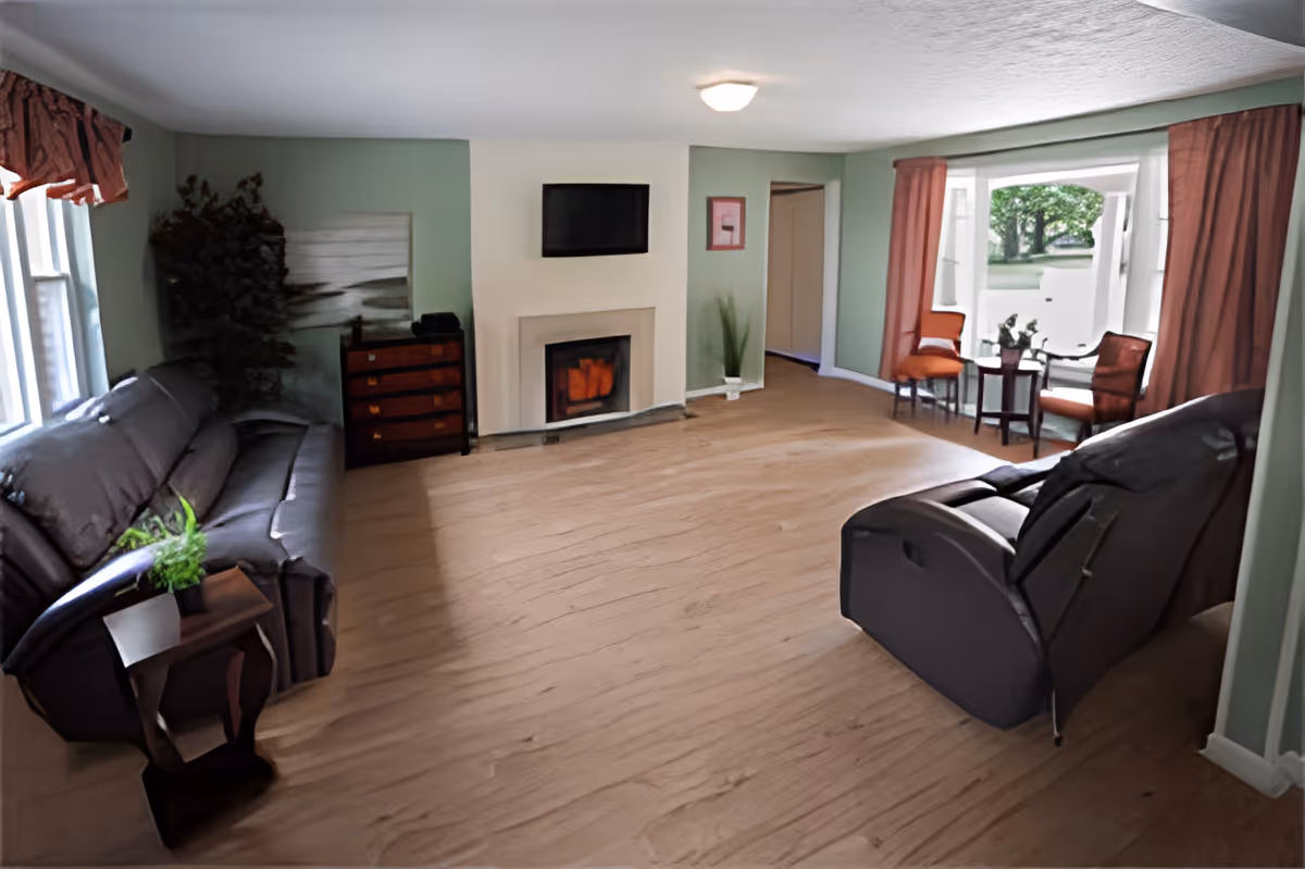 A spacious living room with light green walls and wood flooring. The room features a black leather sofa on the left, a black recliner chair on the right, a small wooden side table with a plant, a fireplace with a mounted TV above it, a chest of drawers, and a bay window area with two orange chairs and a small round table with a plant. The room is well-lit with natural light coming through the windows.