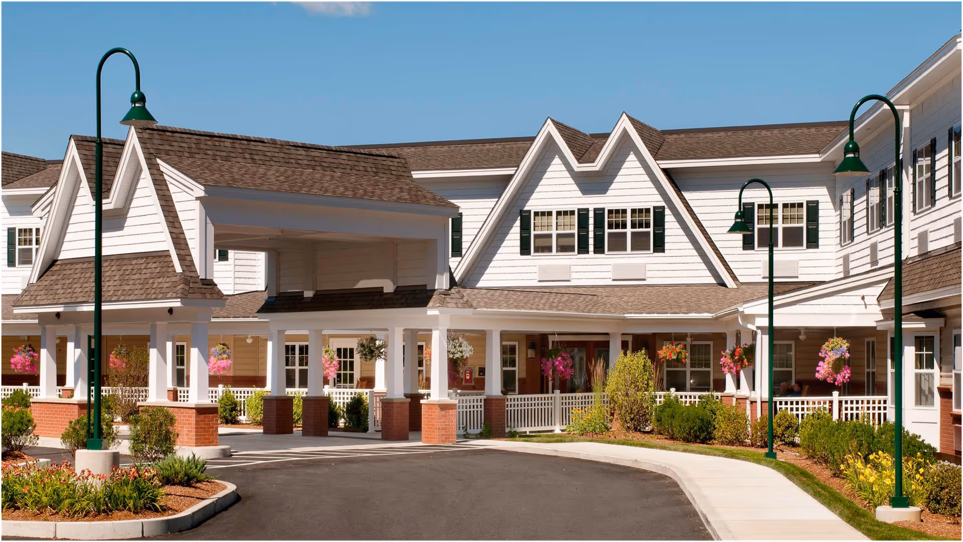 Exterior view of a senior living facility with white siding, brown shingled roofs, green lamp posts, and hanging flower baskets. The building has a covered entrance with brick pillars and a driveway leading up to it.