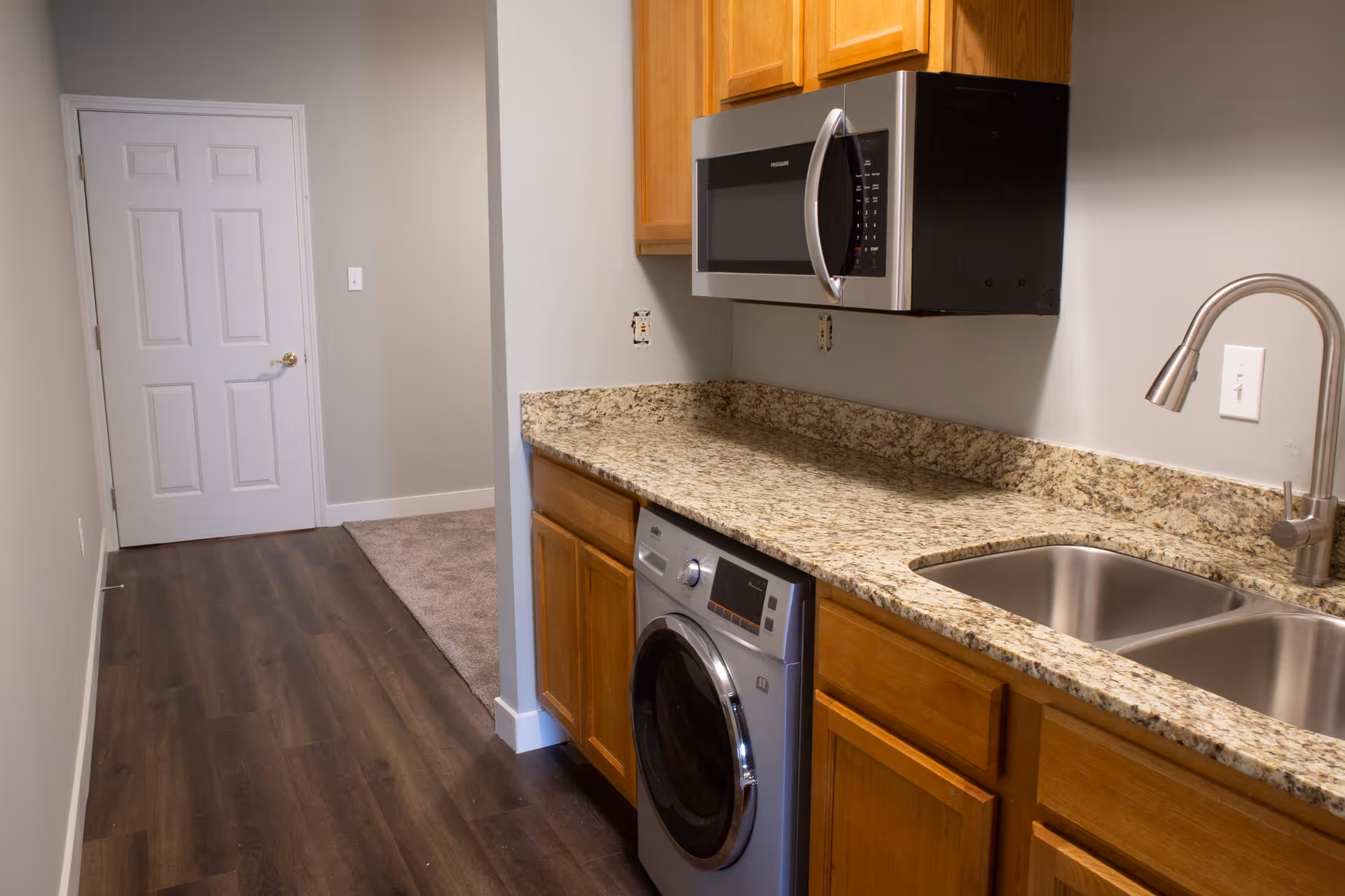 Small kitchenette with granite countertops, stainless double sink, microwave, front-loading washer and wooden cabinets along a dark wood floor.