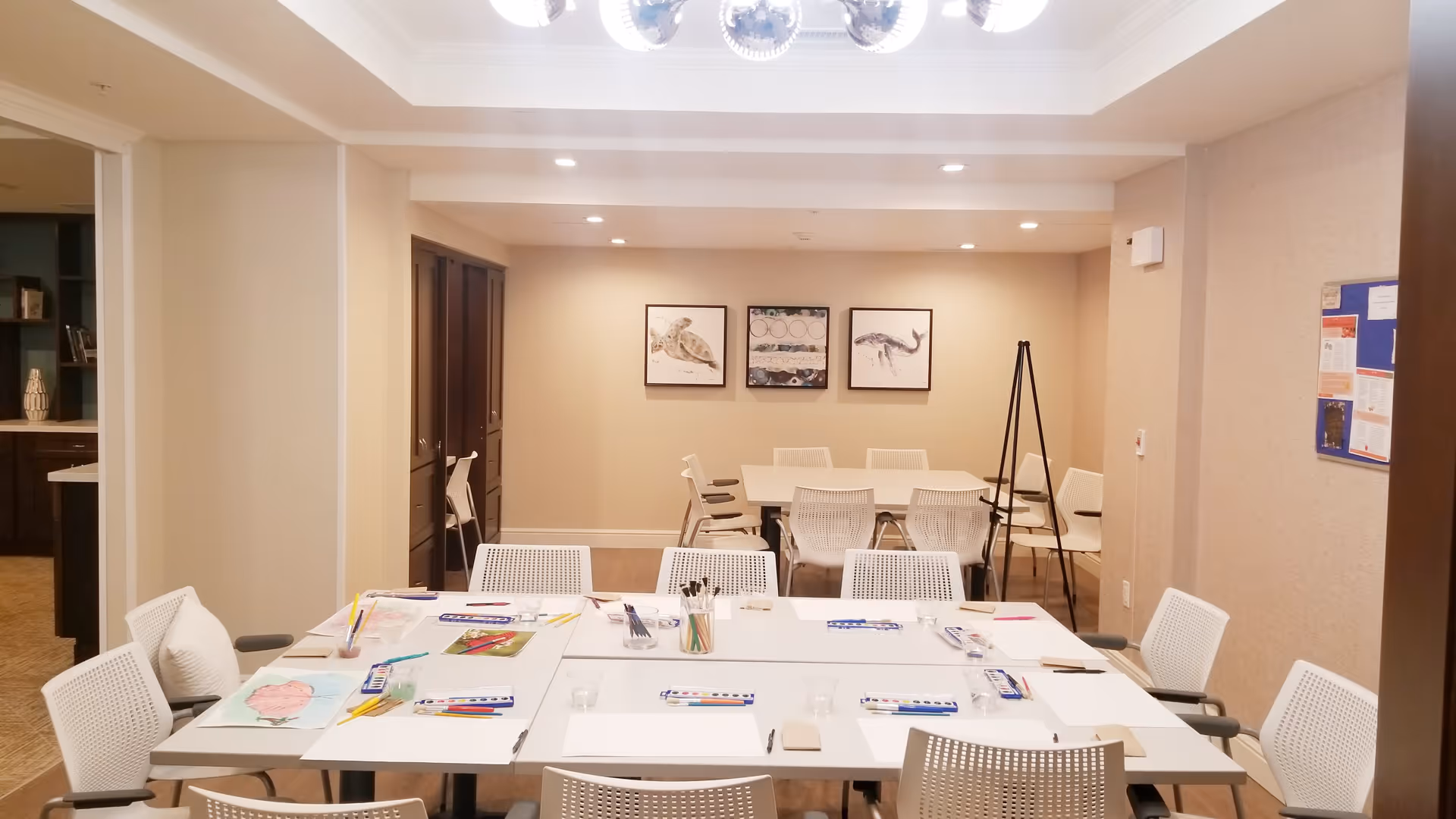 A well-lit room with two white tables arranged for an art or craft activity. The tables have white chairs around them and are set with watercolor paint sets, brushes, pencils, and paper. The walls are beige with three framed pictures of sea creatures and a bulletin board on the right wall. There is a doorway leading to another room with dark wooden cabinets.
