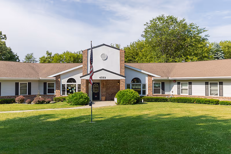 Front exterior view of a single-story senior living facility building with a central entrance, brick and white siding, an American flag on a flagpole in the front lawn, and trees in the background under a partly cloudy sky.
