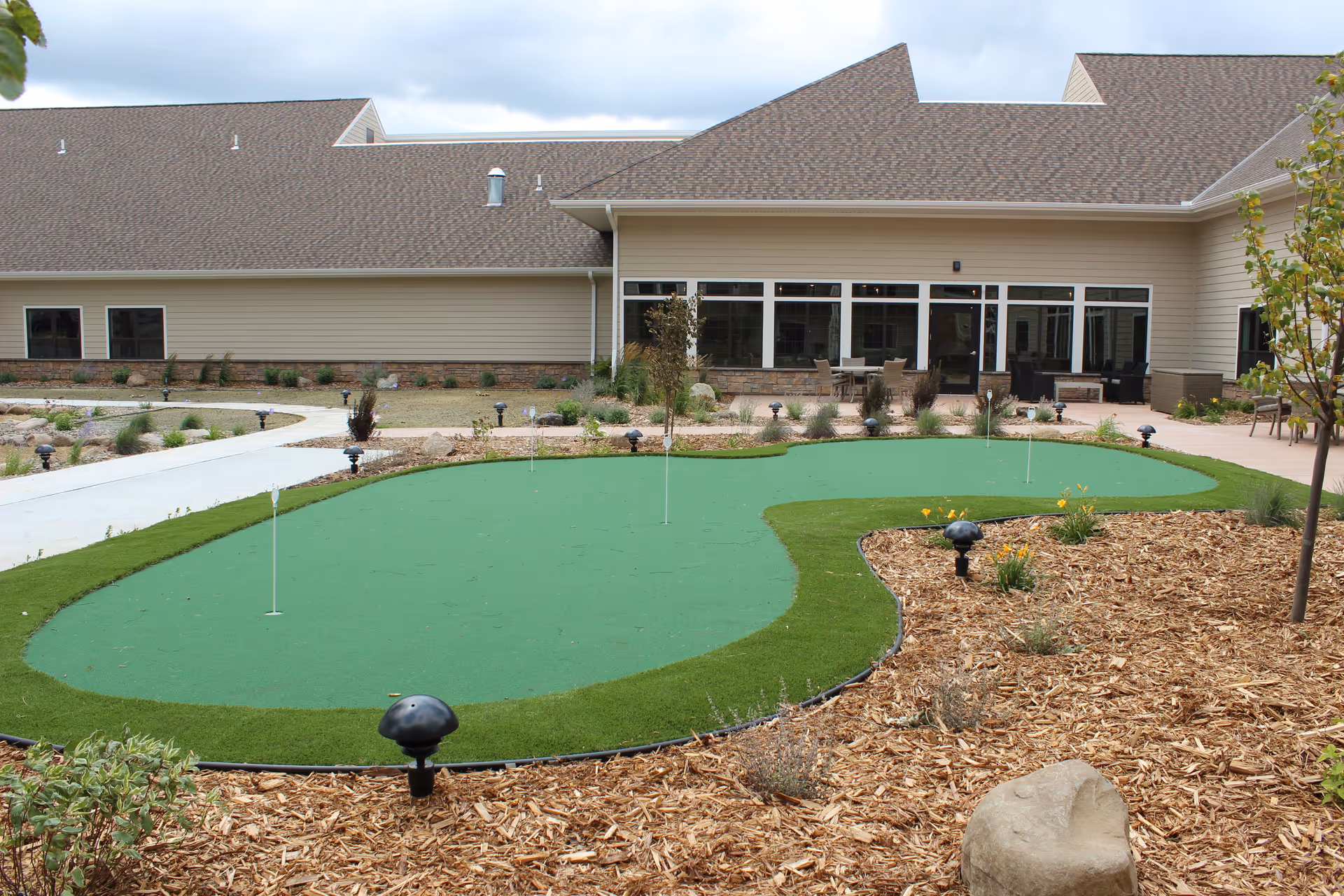 Outdoor courtyard with a small putting green in front of a one-story building with large windows and patio seating.