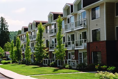 Exterior of a three-story residential building with balconies, landscaped lawn, trees, and a sidewalk.