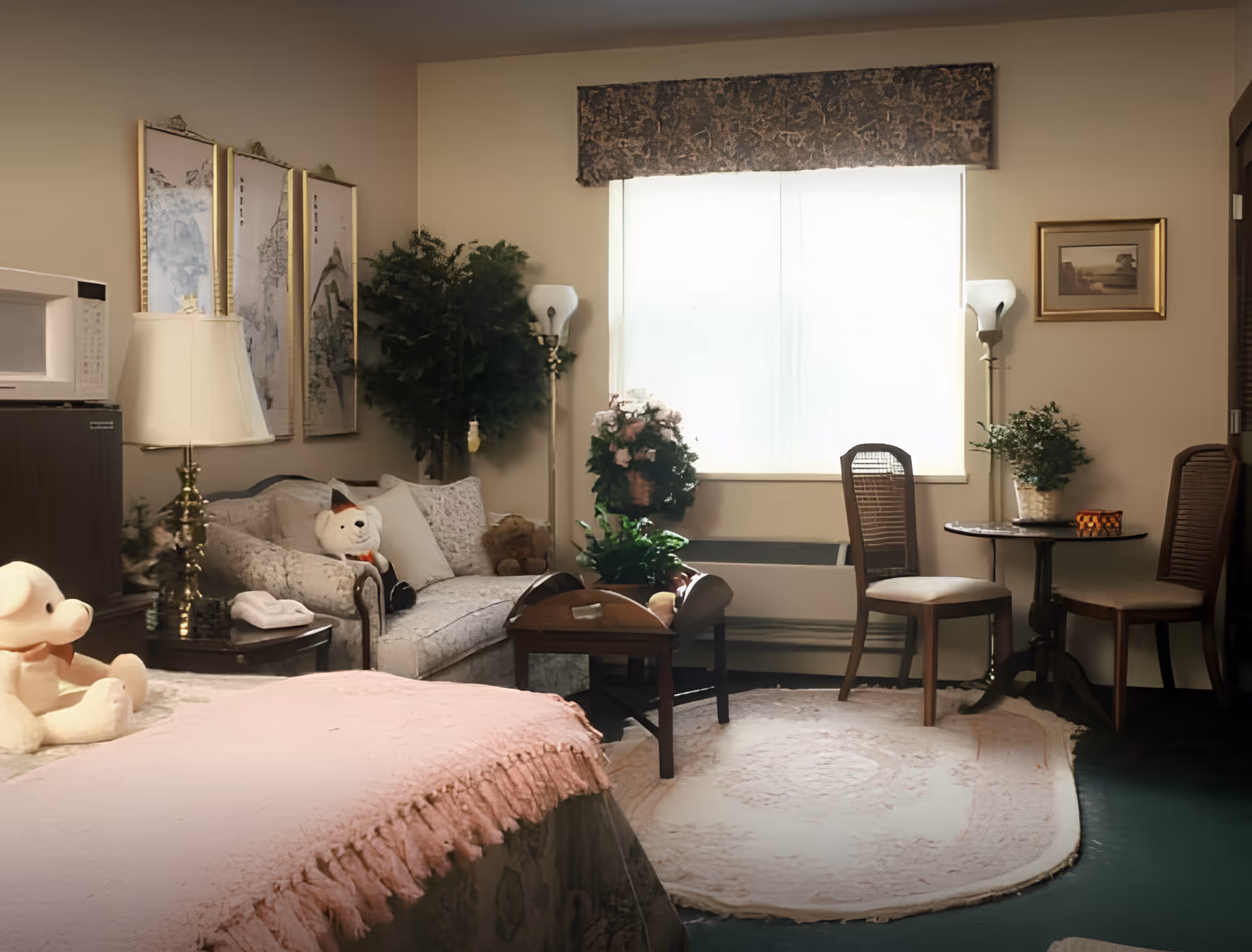 A cozy senior living room with a bed covered in a pink blanket in the foreground, a small sofa with stuffed animals, a side table with a lamp, a coffee table, two wooden chairs around a small round table, several potted plants, and framed artwork on the walls. A large window with a valance allows natural light into the room.