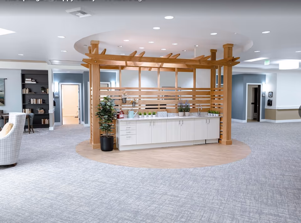 Open senior-living common room with a central wooden pergola-style kitchenette/serving counter and seating areas.