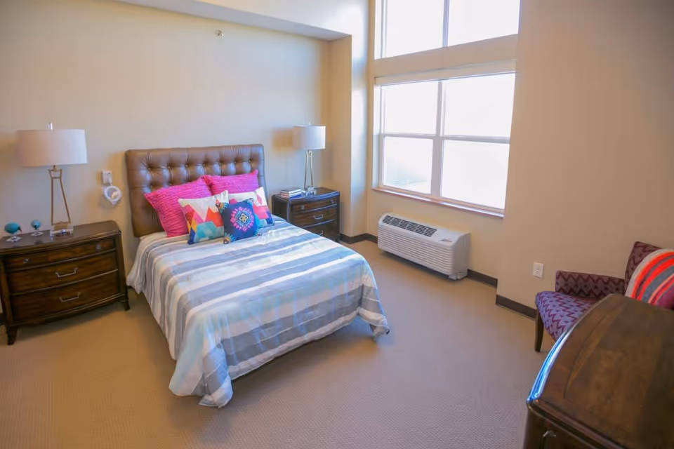 A neatly furnished bedroom with a tufted headboard bed, nightstands with lamps, a chair and a large window letting in natural light.