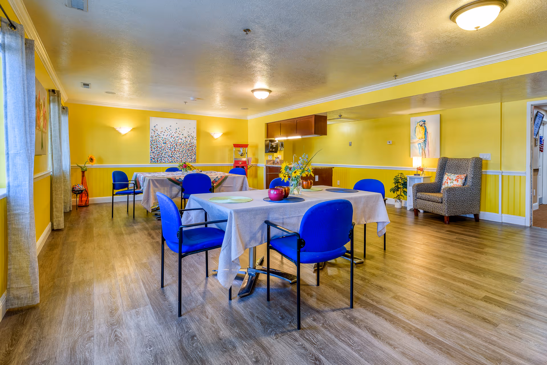Bright communal dining room with yellow walls, tables set with cloths and blue chairs and a small sitting area.