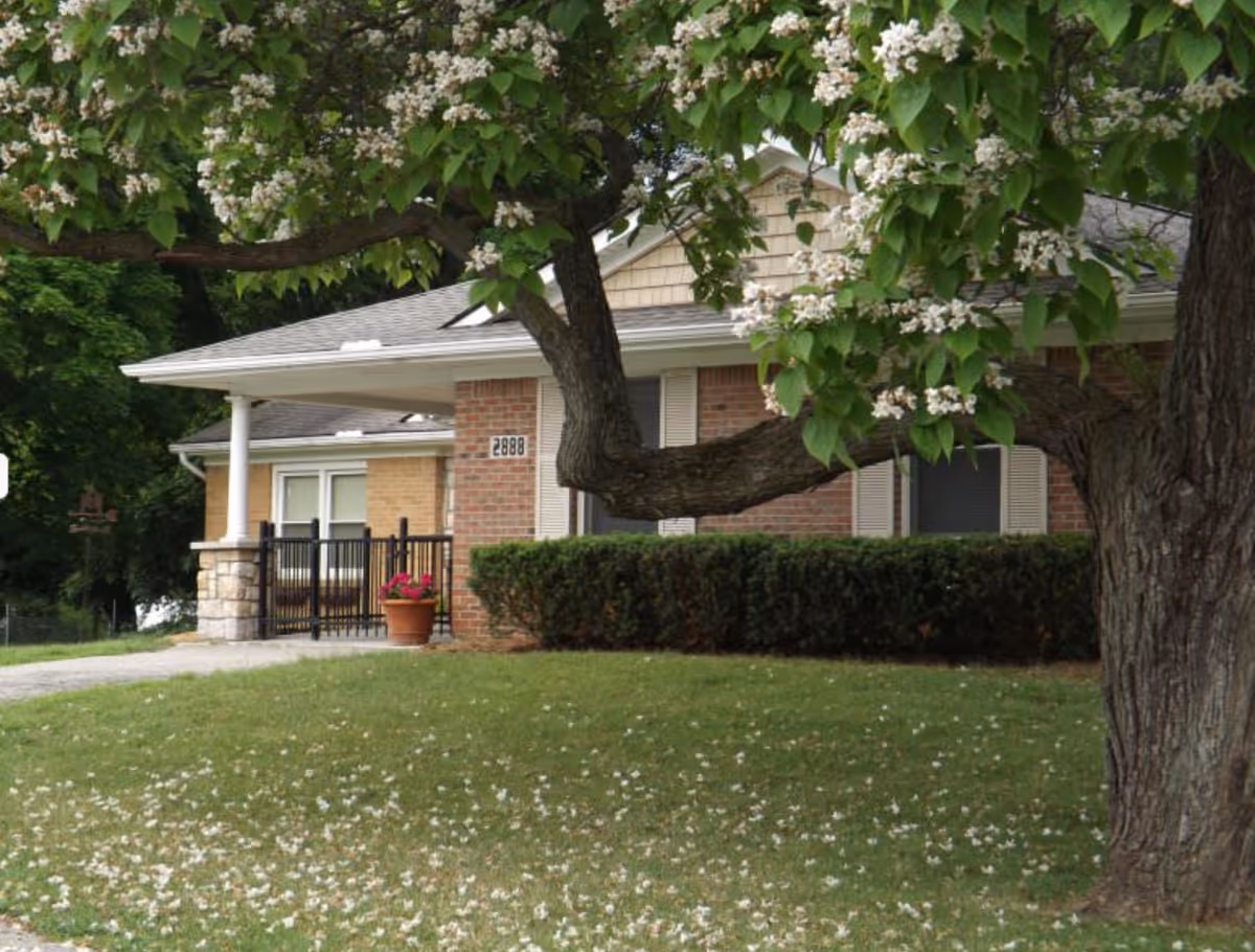 Exterior view of a single-story brick building with a covered porch, surrounded by green grass and trees with white blossoms in the foreground.