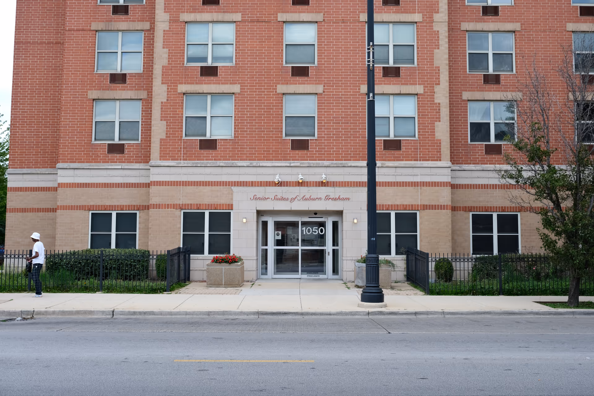 Front exterior view of a multi-story brick building with the entrance labeled Senior Suites of Auburn Gresham and the address number 1050 above the door. There is a sidewalk, a street lamp, some greenery, and a person walking on the left side of the image.