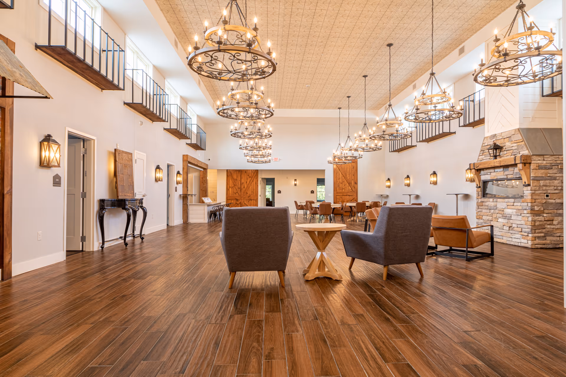 Spacious and well-lit common area with wooden flooring, multiple round chandeliers hanging from the ceiling, and a stone fireplace on the right. The room features several seating arrangements including gray and brown chairs around small wooden tables. There are large wooden barn-style doors at the far end and multiple small balconies along the upper walls.