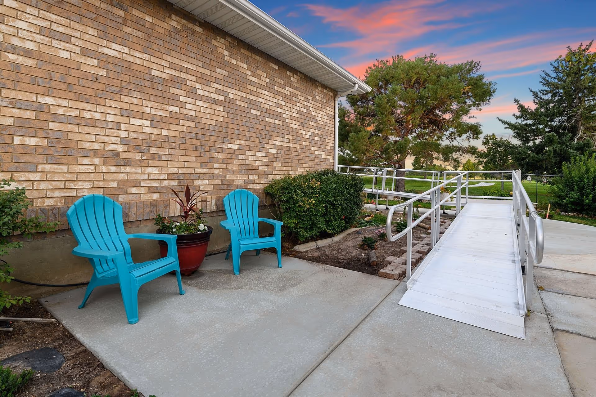 Outdoor patio area next to a brick building wall with two turquoise plastic chairs and a large potted plant. There is a metal wheelchair ramp leading to a grassy area with trees under a colorful sunset sky.