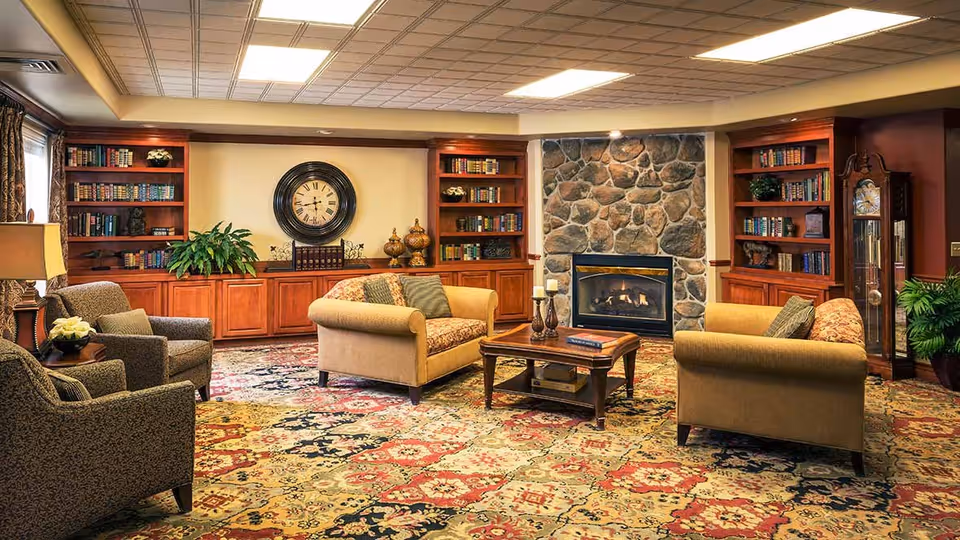 A cozy living room with two beige sofas and two armchairs arranged around a wooden coffee table. The room features a stone fireplace with a fire burning, built-in wooden bookshelves filled with books and decorative items, a large wall clock, a grandfather clock, and a patterned carpet covering the floor.