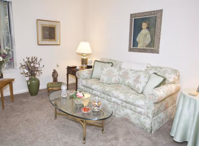 Carpeted living room with a floral-patterned sofa, glass coffee table, side tables with a lamp and decorations, and framed artwork on the walls.