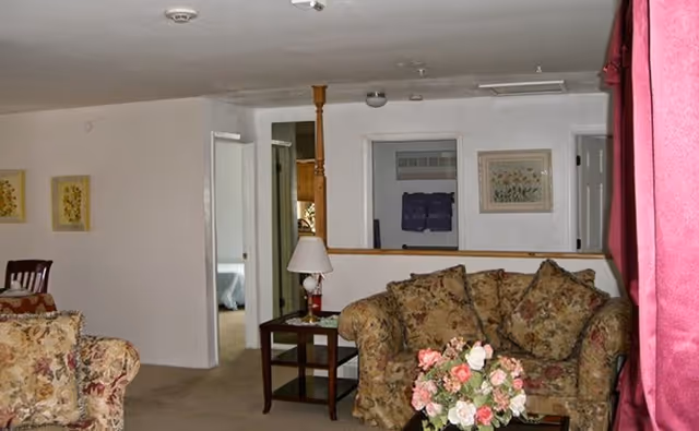 Interior view of a living room area with floral patterned sofas, a wooden side table with a lamp, and a bouquet of pink and white flowers on a table in the foreground. The room has white walls with framed artwork and an open doorway leading to another room with a bed visible.
