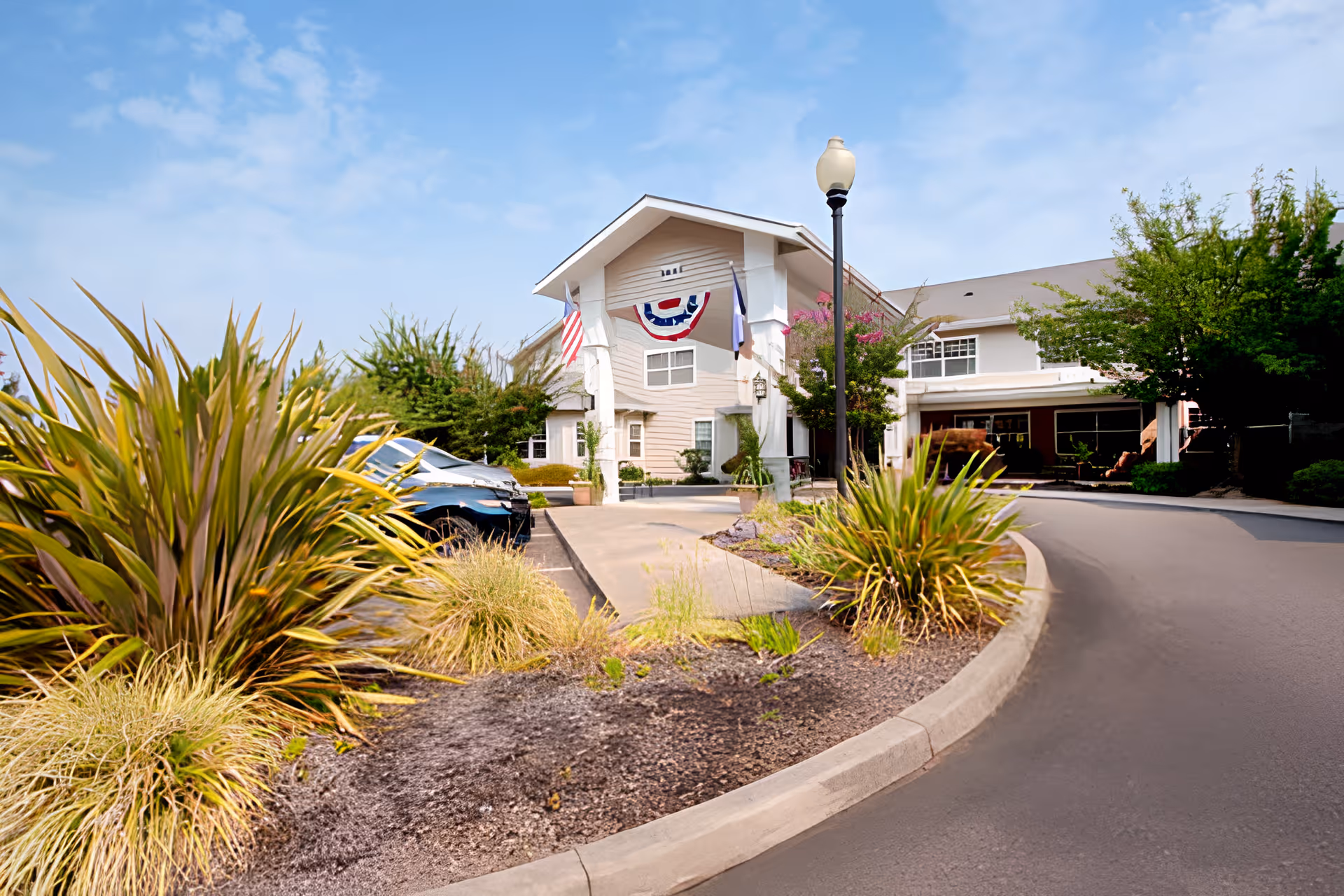 Entrance and front facade of a senior living building with a curved driveway, landscaped islands, flags, and decorative bunting.