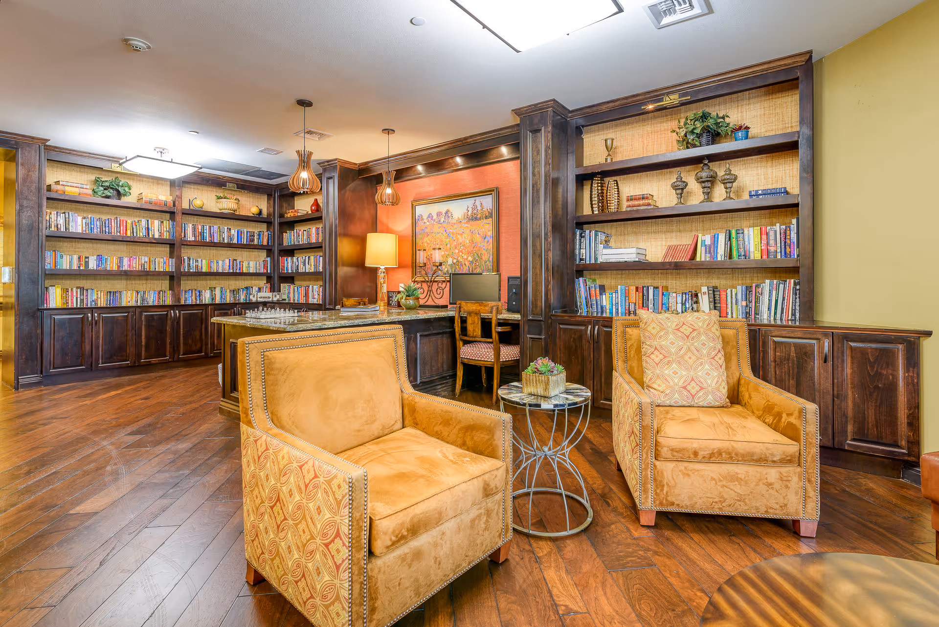 A cozy library or reading room with wooden bookshelves filled with books and decorative items. Two mustard-colored armchairs with patterned sides and a small round table with a plant are in the foreground. A desk with a computer and chair is set against a wall with a framed painting and a table lamp. The room has warm lighting and wooden flooring.