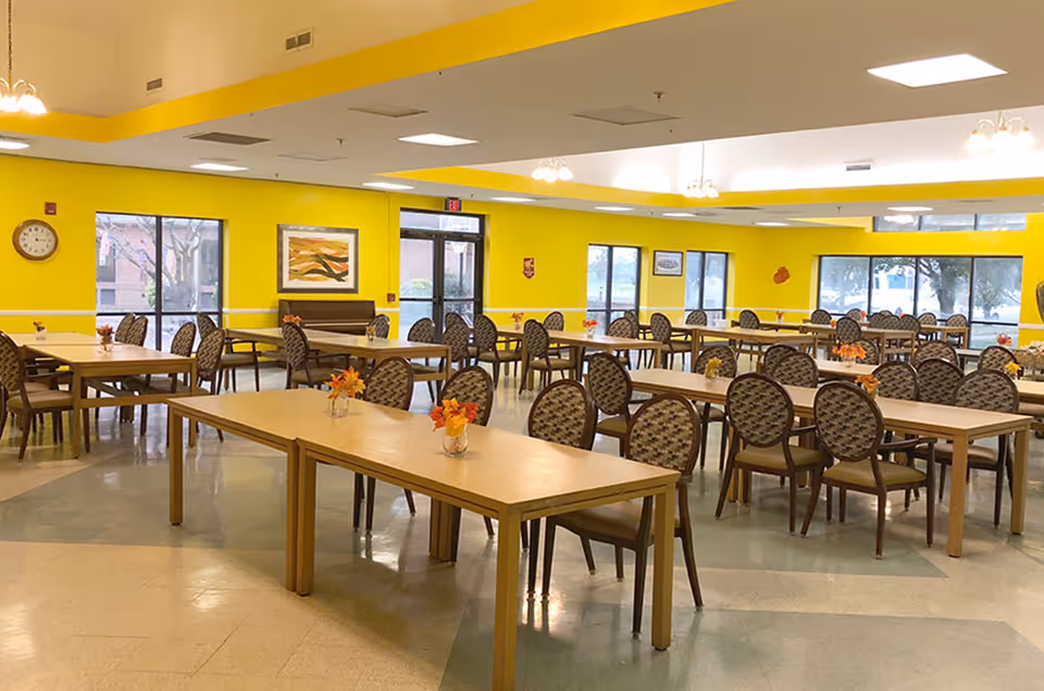A large dining room with multiple wooden tables and patterned chairs arranged neatly. The walls are painted bright yellow with several windows letting in natural light. Small flower arrangements are placed on each table. There is a clock on the wall and framed artwork, with ceiling lights providing additional illumination.