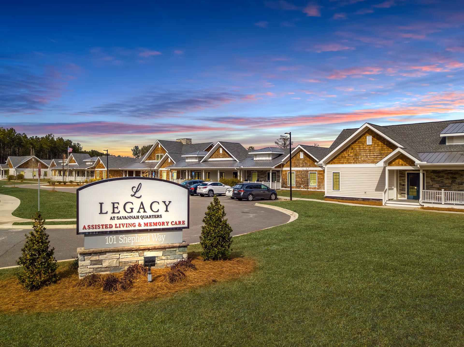 Front view of the Legacy at Savannah Quarters assisted living and memory care campus with a prominent entrance sign, parking area, and single-story buildings under a colorful sky.