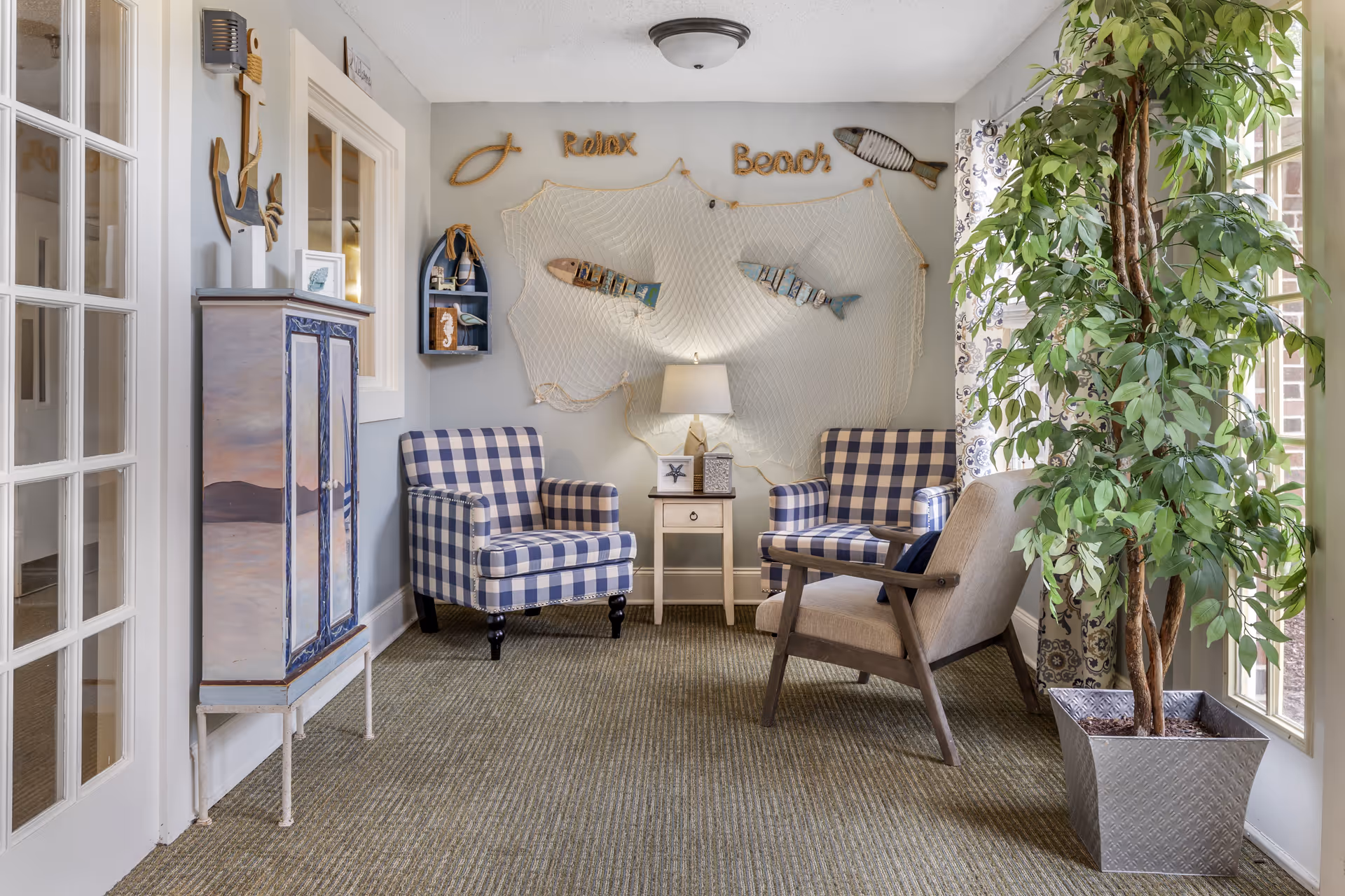 A cozy sitting area with two blue and white checkered armchairs and one beige armchair arranged around a small wooden side table with a lamp. The wall behind features nautical-themed decorations including a fishing net, wooden fish, and the words 'Relax' and 'Beach'. A tall potted plant is placed near a window with patterned curtains, and a painted cabinet with an anchor decoration is on the left side.