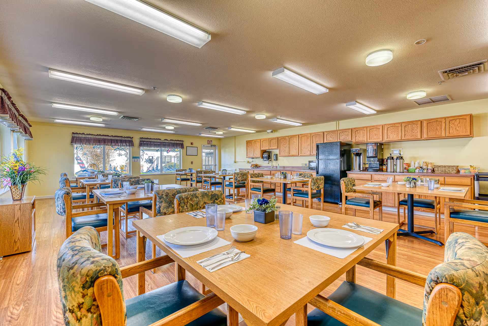 A bright and spacious dining room in a retirement community with multiple wooden tables and chairs arranged neatly. Each table is set with white plates, bowls, utensils, and glasses. The room has large windows with valances, wooden cabinetry along one wall, and various beverage dispensers and condiments on the counter. The floor is wooden, and the ceiling has multiple fluorescent lights.