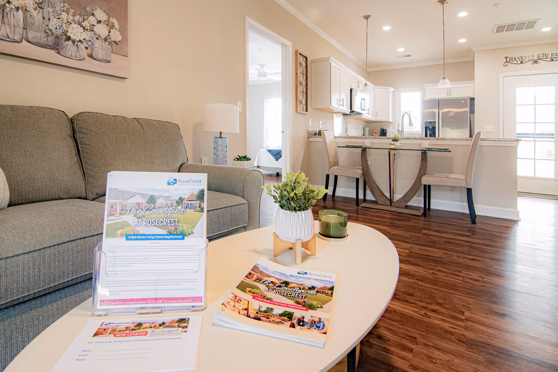 A bright and modern living area in a senior living facility with a gray sofa, a white oval coffee table holding brochures and a small plant, and a view into a kitchen with a glass dining table and two beige chairs. The kitchen features white cabinets, stainless steel appliances, and wood flooring. A bedroom is visible through an open doorway.