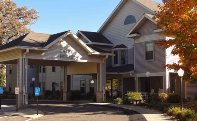 Exterior view of a senior living facility building named Care House with a covered entrance, surrounded by trees with autumn foliage and a paved driveway with handicap parking signs.