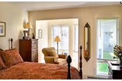 A cozy bedroom with a large bed covered in a patterned red bedspread, a wooden dresser with a lamp and framed picture, a comfortable armchair near a window, and a tall mirror on the wall. The room has light-colored walls and a window letting in natural light.