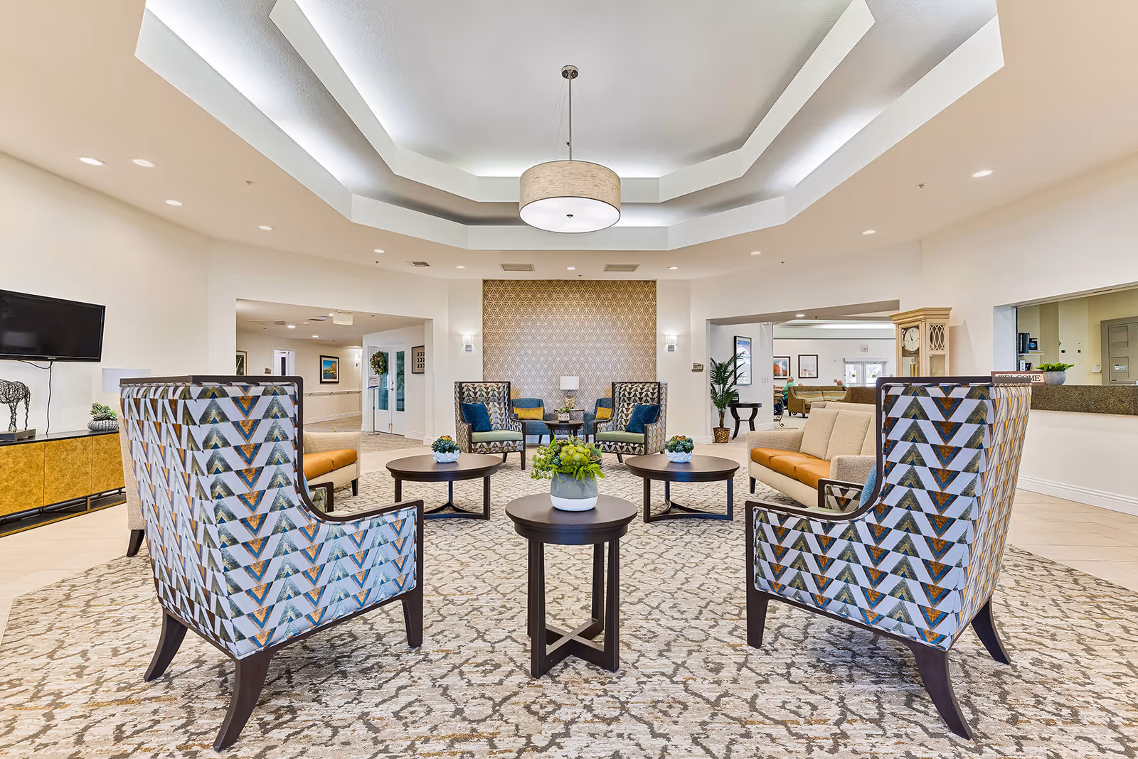 Bright senior living lobby with patterned armchairs, sofas, round coffee tables and a decorative recessed ceiling light.