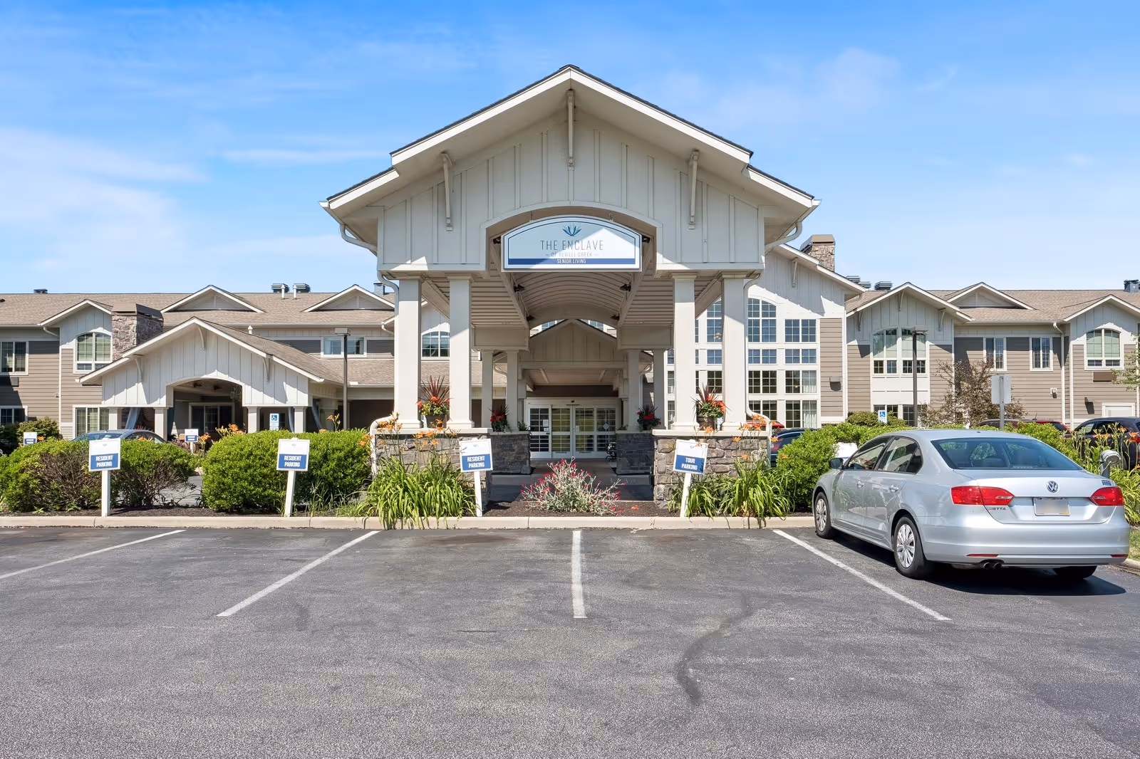 Front exterior view of The Enclave of Newell Creek senior living facility with a covered entrance, parking spaces, and a silver car parked on the right side. The building has multiple windows and a beige and white color scheme under a clear blue sky.
