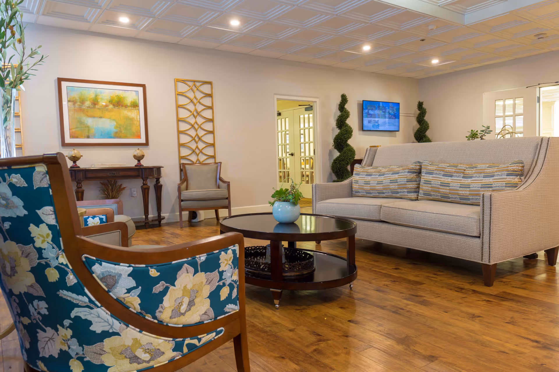 A cozy senior living facility lounge area with a beige sofa adorned with striped cushions, a round dark wood coffee table with a small plant, and two floral-patterned armchairs. The room has wooden flooring, a decorative wall art piece, a small wooden console table with two small globes, and a flat-screen TV mounted on the wall displaying a welcome message. There are also two spiral-shaped topiary plants near the TV and a doorway leading to another room with glass-paneled doors.