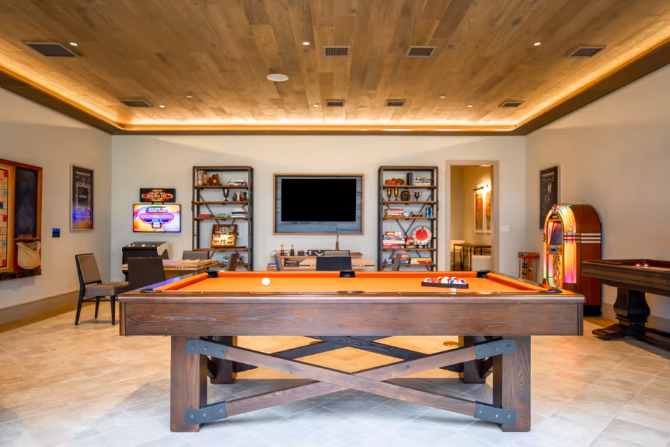 A recreational room with a wooden pool table featuring an orange felt surface in the center. Behind the pool table are two metal and wood bookshelves filled with books and decorative items, and a large flat-screen TV mounted on the wall between them. To the left, there is a small table with chairs and a wall-mounted game board. On the right side, a vintage-style jukebox and another game table are visible. The room has a wooden ceiling with recessed lighting and light-colored tiled flooring.