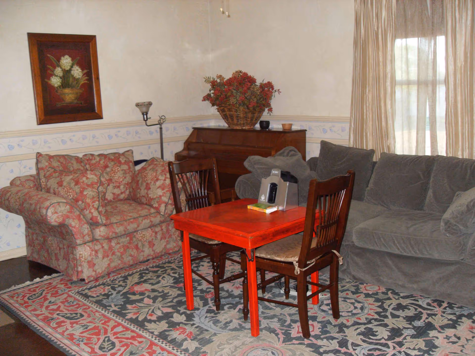 A cozy living room with a floral patterned armchair, a gray sofa, a red wooden table with two wooden chairs, a floor lamp, a piano with a basket of red flowers on top, and a framed floral painting on the wall. The room has beige walls with a decorative border and a large window with beige curtains.